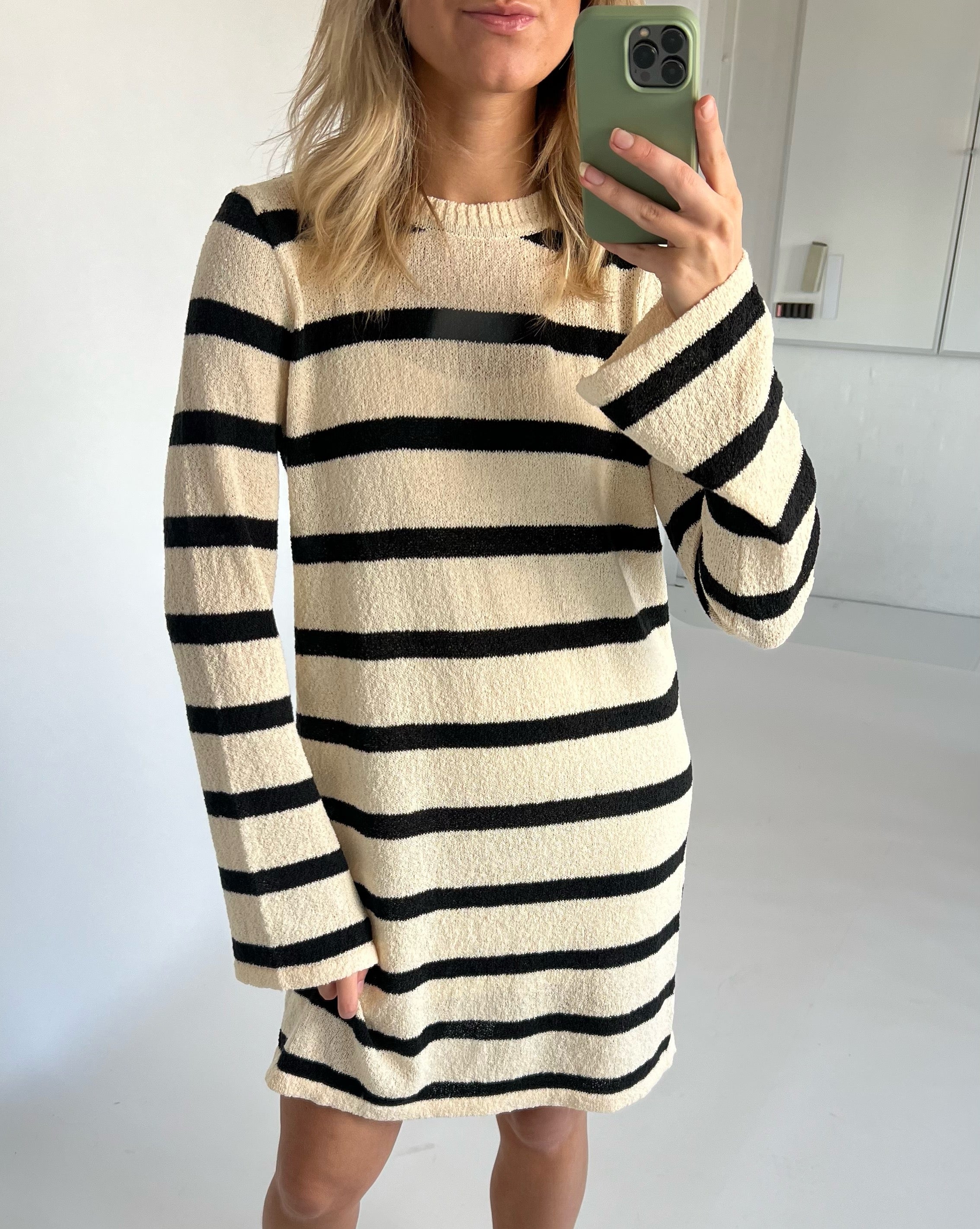 Kvinde i beige og sort-stribet kjole tager selfie med mobiltelefon i et hvidt rum.