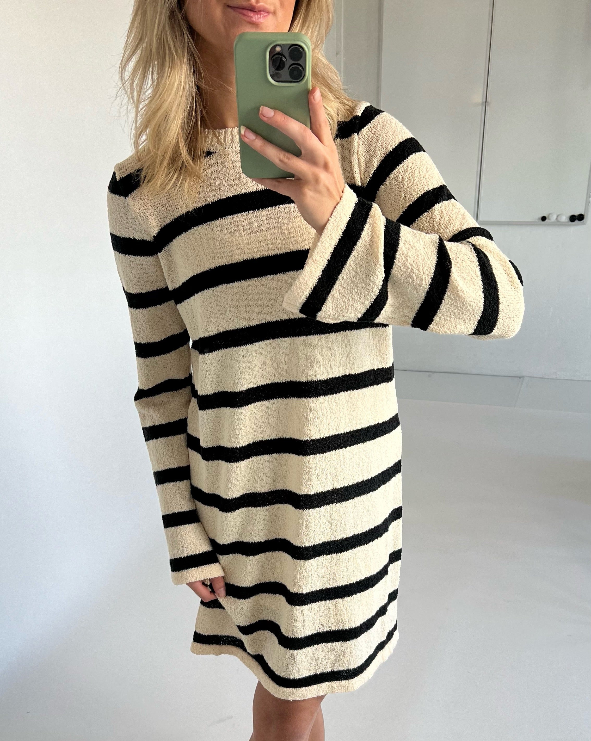 En person i en beige og sort stribet kjole tager et spejlbillede med en grøn telefon i et lyst rum.
