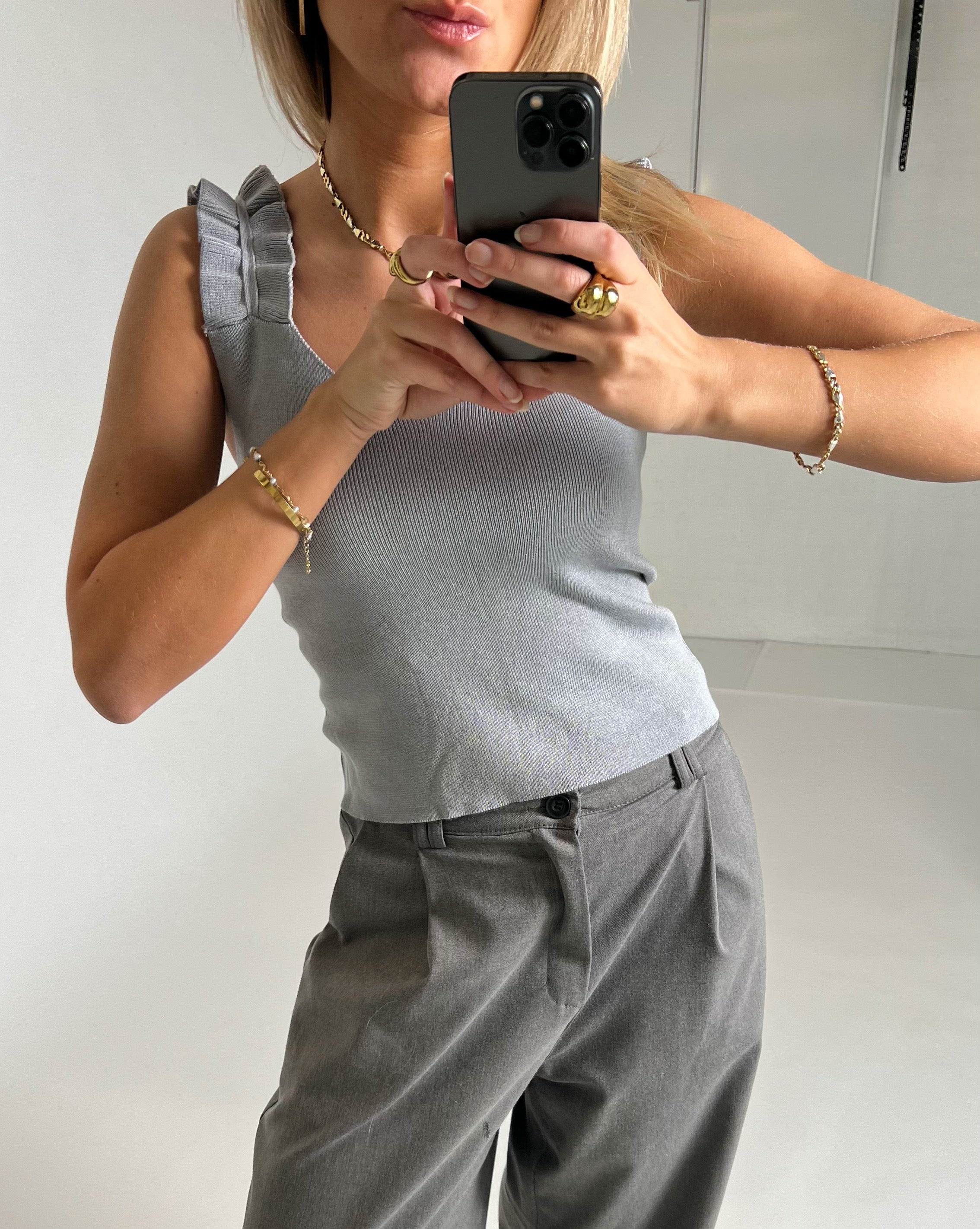En person i grå top og bukser tager et spejlbillede med en smartphone. Personen bærer gyldne smykker, og omgivelserne er minimalistiske og lyse.