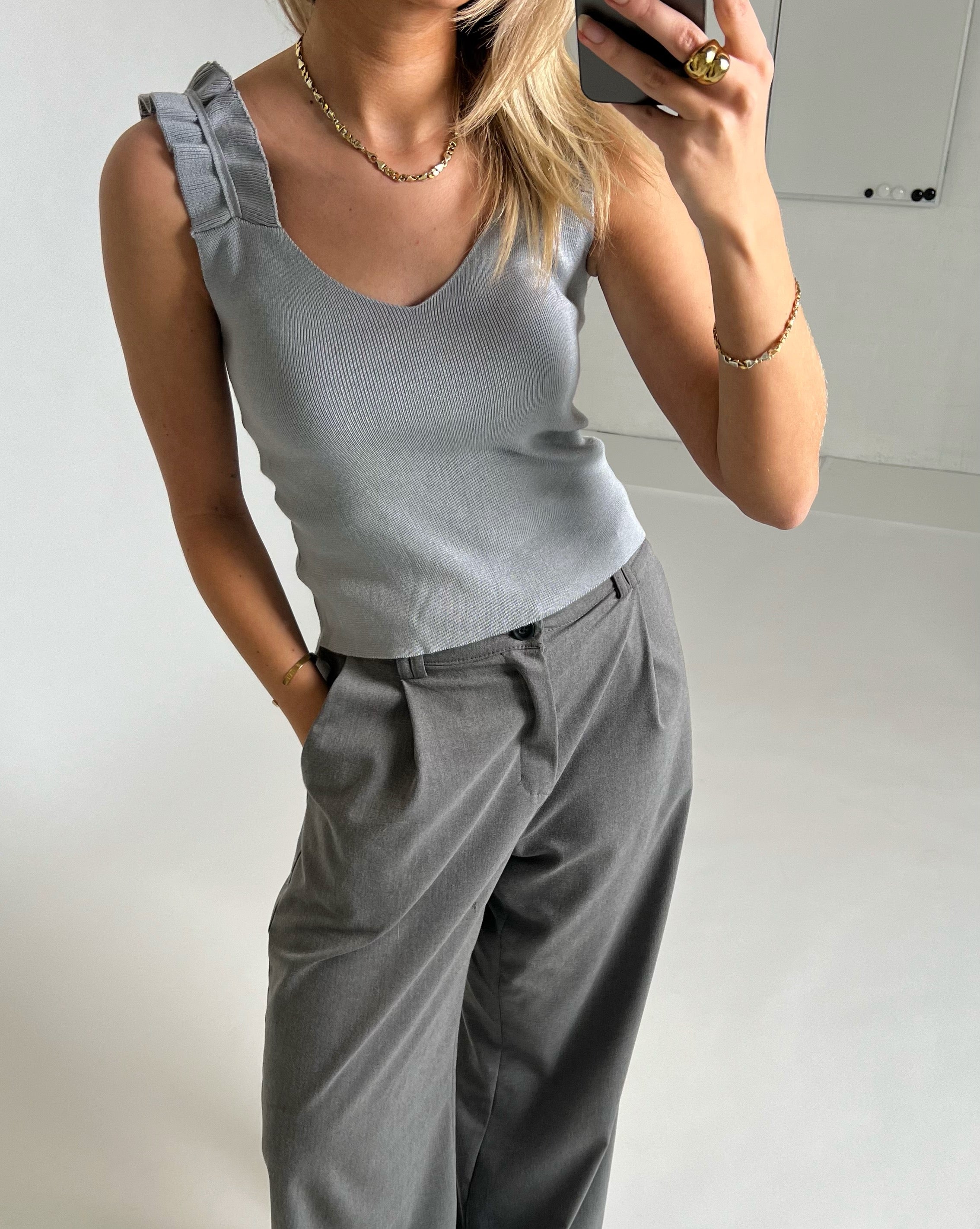 En person iført en lysegrå top og grå bukser tager en selfie. Personen står i et minimalistisk, lyst rum og bærer smykker.