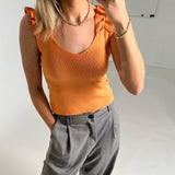 En person iført en orange top med flæser og grå bukser tager et selfie i en hvidmalet indendørs setting.