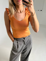 En person iført en orange top med flæser og grå bukser tager et selfie i en hvidmalet indendørs setting.
