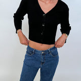 Kvinde bærer sort cardigan med knapper og blå jeans, står i afslappet pose med hænderne i lommerne, mod en simpel hvid baggrund.