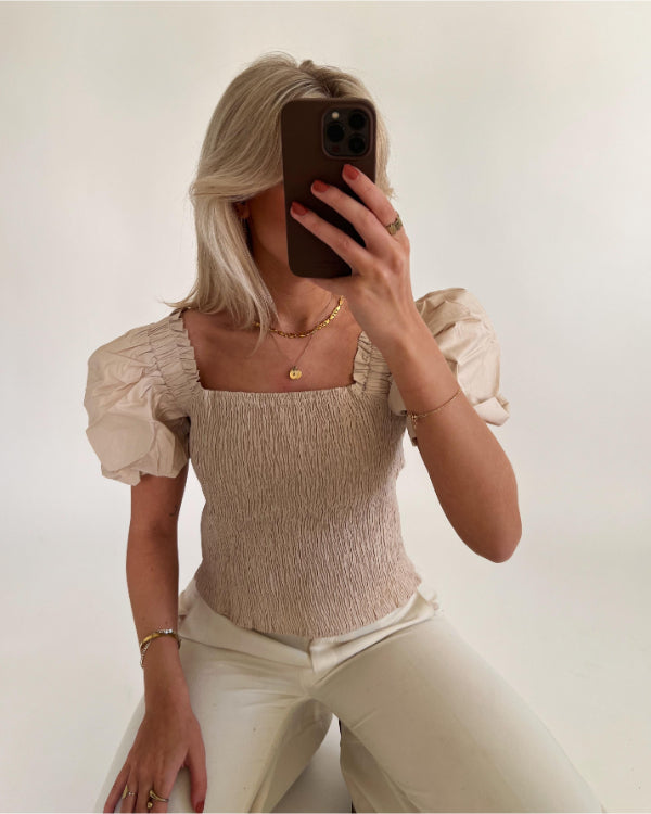 En person med lyst hår tager en selfie med en smartphone i et hvidt rum. De bærer en lys bluse med pufærmer og hvide bukser.