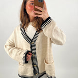 Person tager et selfie i spejl. Personen har en beige cardigan med sorte kanter og knapper, står foran en ensfarvet hvid baggrund. Hun holder en telefon med brun beskyttelse.