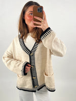 Person tager et selfie i spejl. Personen har en beige cardigan med sorte kanter og knapper, står foran en ensfarvet hvid baggrund. Hun holder en telefon med brun beskyttelse.