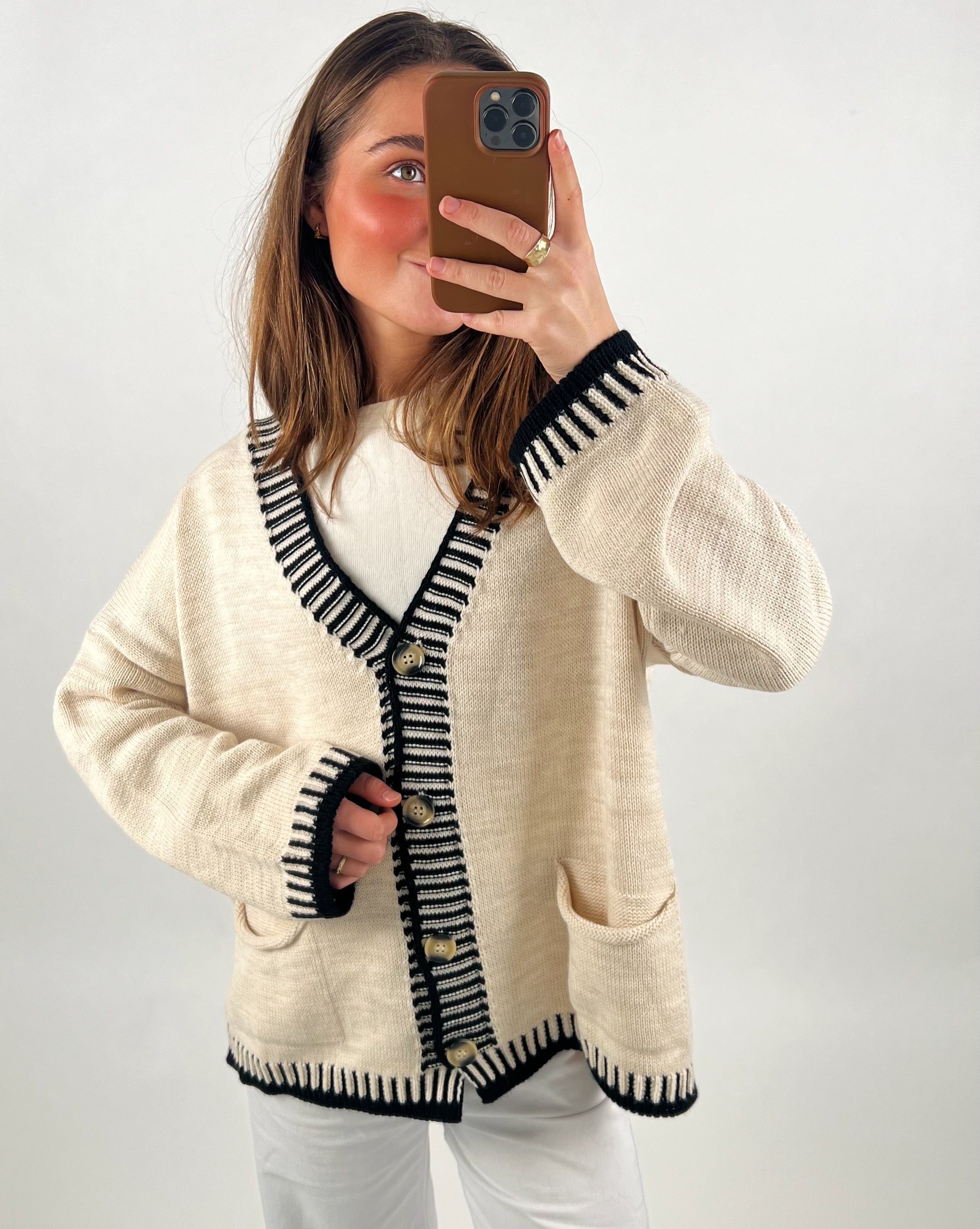 Person tager et selfie i spejl. Personen har en beige cardigan med sorte kanter og knapper, står foran en ensfarvet hvid baggrund. Hun holder en telefon med brun beskyttelse.