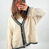 En person tager et spejlbillede med en smartphone, iført en beige cardigan med sorte striber og knapper. Indendørs og tydeligt lys. Ingen tekst til stede.