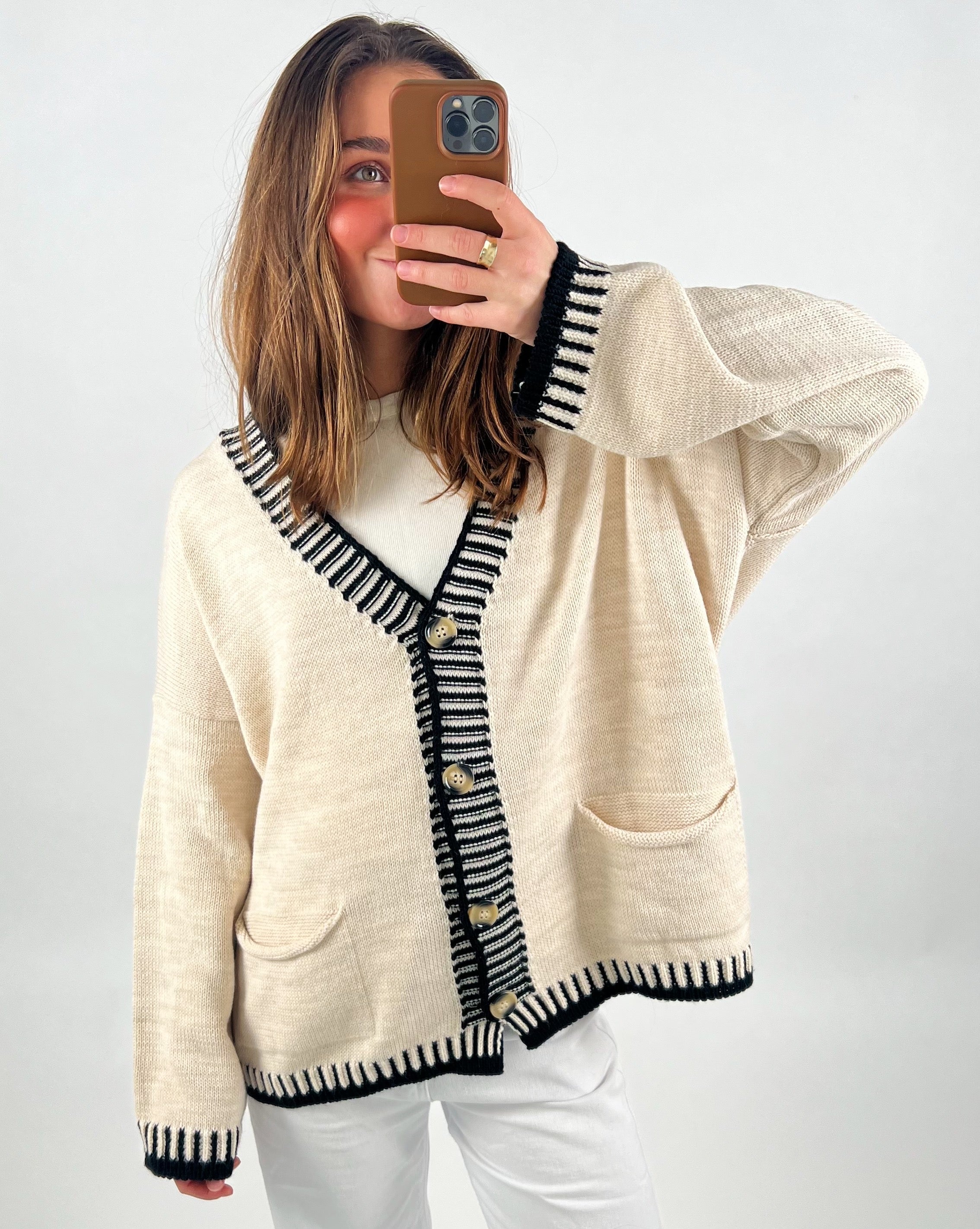 En person tager et spejlbillede med en smartphone, iført en beige cardigan med sorte striber og knapper. Indendørs og tydeligt lys. Ingen tekst til stede.