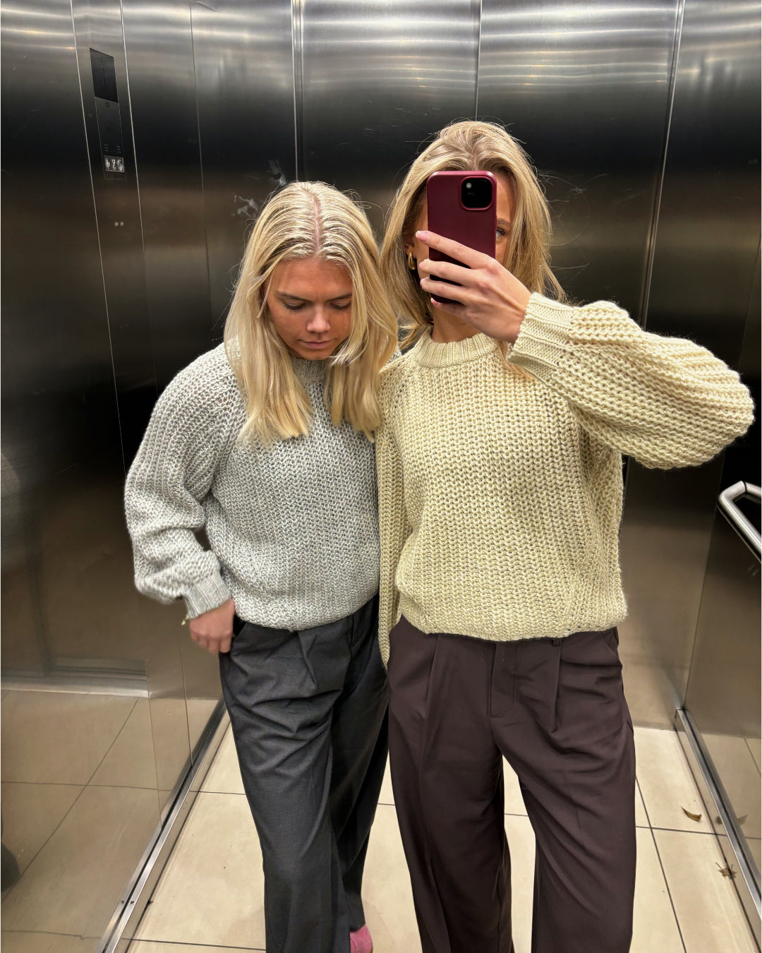 To personer i striktrøjer står i en elevator med et spejl. Den ene tager et selfie med en smartphone. Elevatorens blanke metalvægge og flisegulv ses omkring dem.