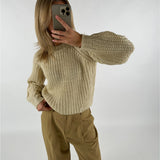 En person tager en selfie i spejlet iført en strikket, cremefarvet sweater med lange ærmer og beige bukser i et minimalistisk rum.