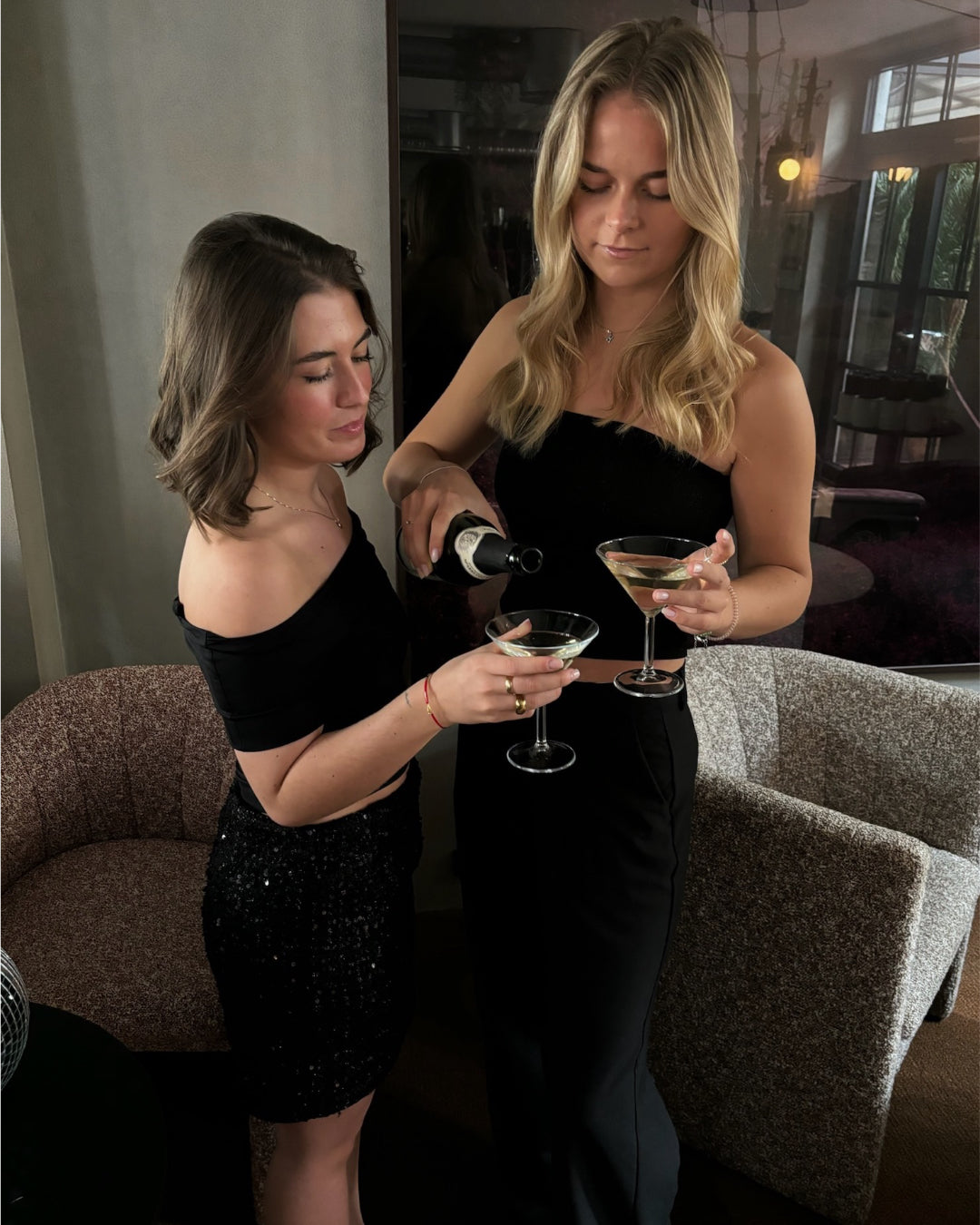 To kvinder i sorte kjoler hælder champagne i glas, mens de står ved siden af en beige lænestol i et moderne rum med store vinduer.