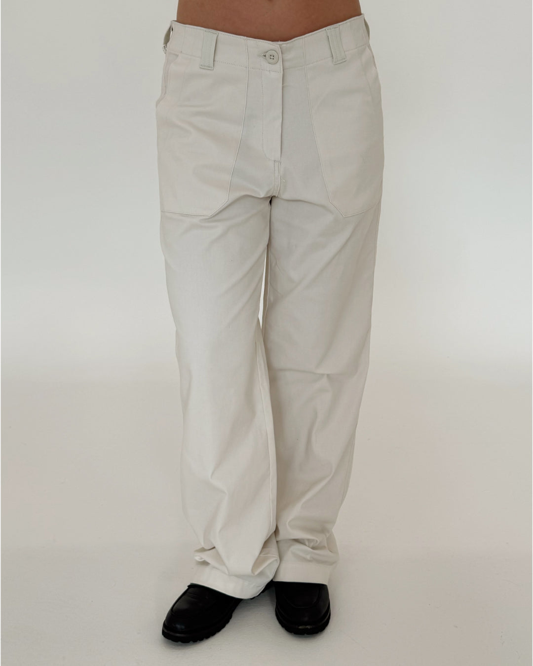 Bukser, iført af en person, er lyse og vide, parret med sorte sko. Baggrunden er enkel og hvid, hvilket fremhæver det minimalistiske look.