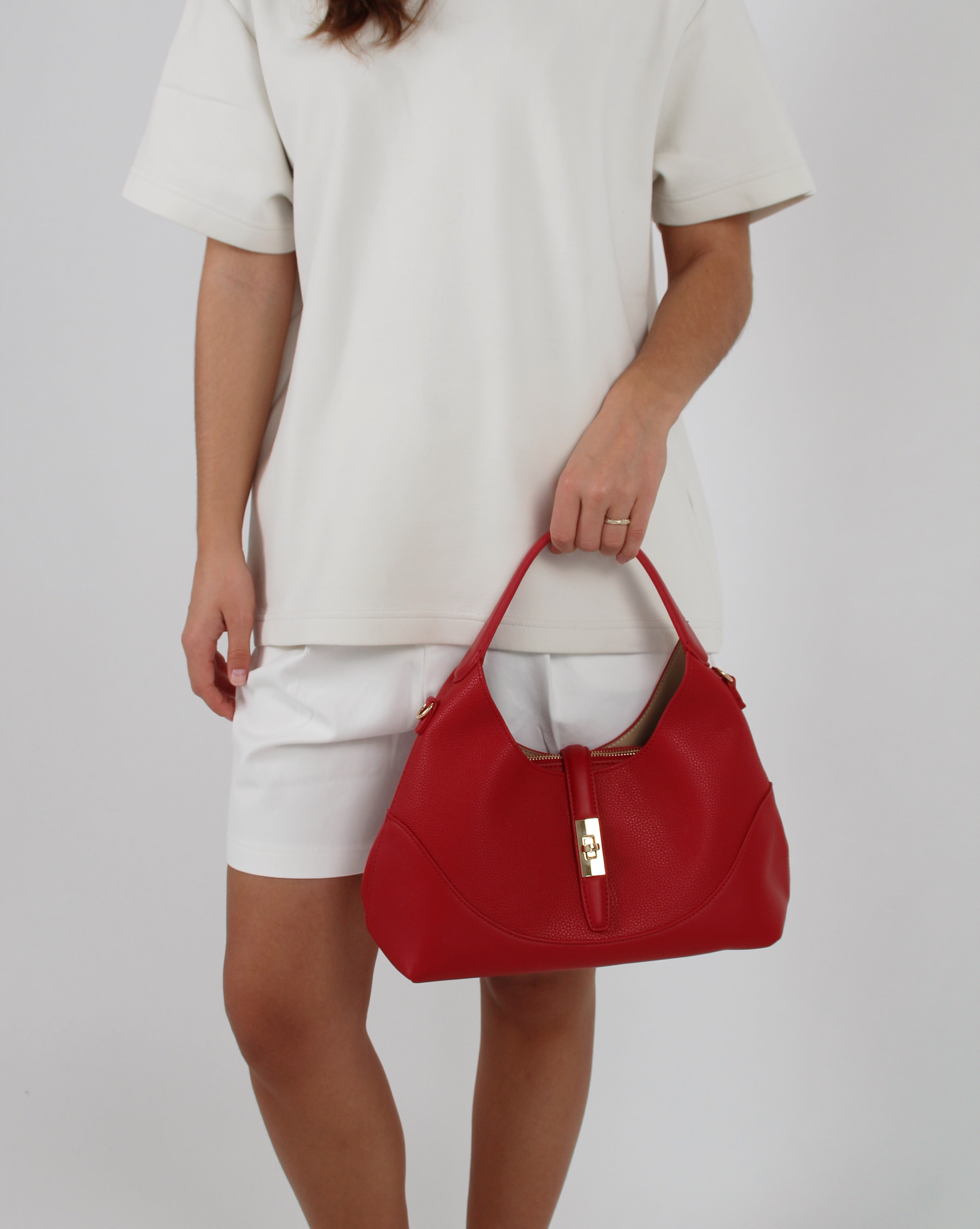 Rød håndtaske bæres af en person i hvide shorts og T-shirt mod en grå baggrund. Tasken har en gylden lås og kort håndtag.