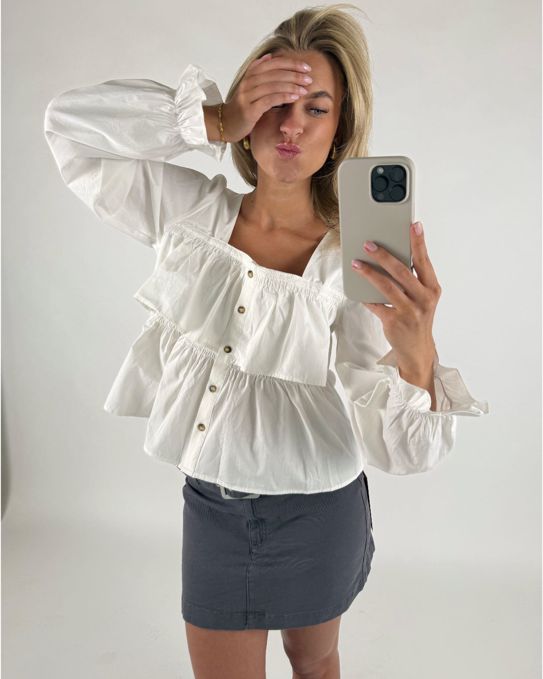 Person tager en selfie iført en hvid, detaljeret bluse og mørkt nederdel i et lyst rum. Personen har hånden over øjnene og laver et kysseansigt, mens mobilen fanger billedet.