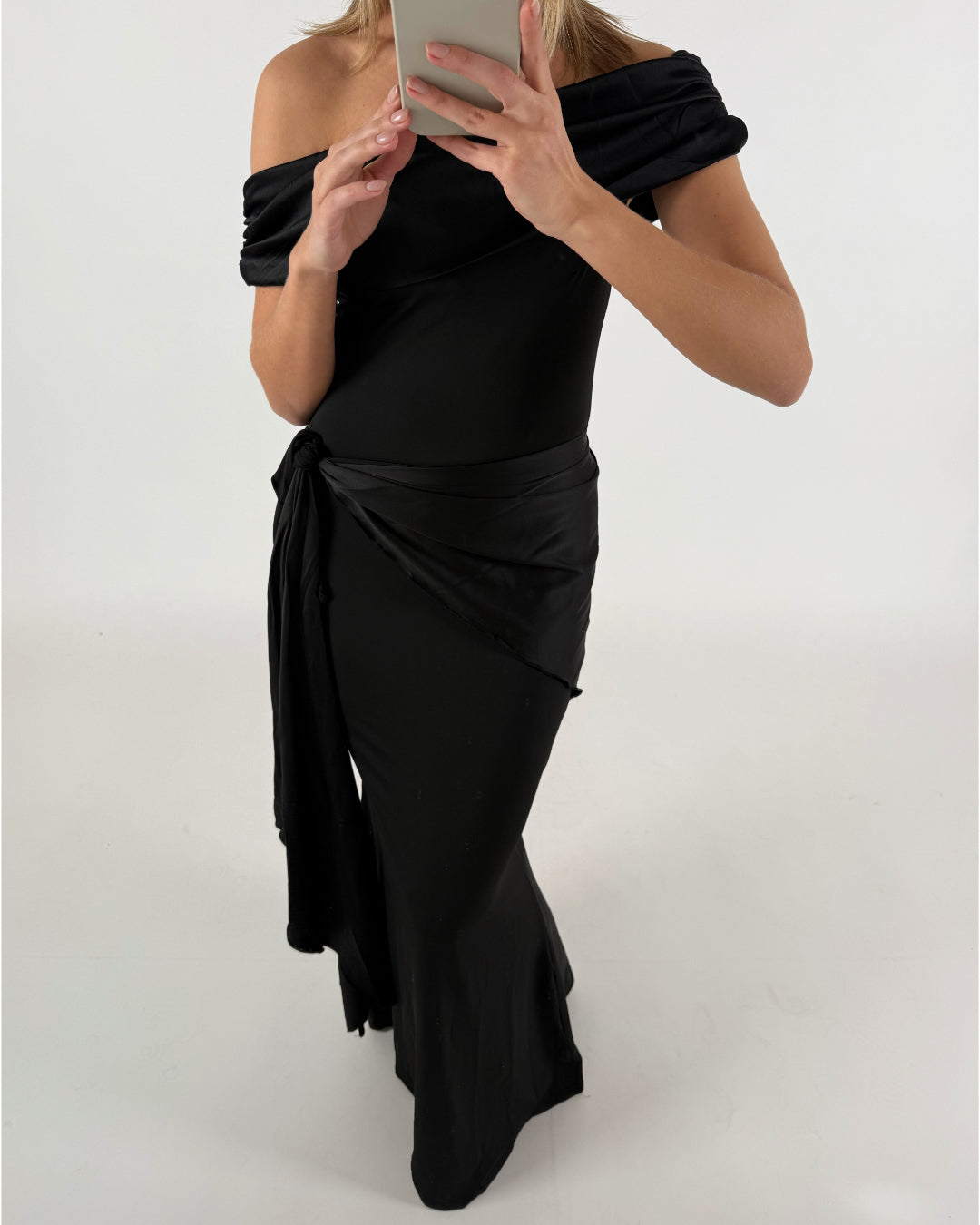 En person iført en sort off-shoulder kjole tager et spejlfoto i et minimalistisk rum.