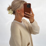 En person med lyst hår i en opsat frisure med en blomsterformet hårklemme tager et selfie med en mobiltelefon. Personen bærer en lys cardigan i et neutralt rum.