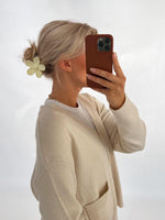 En person med lyst hår i en opsat frisure med en blomsterformet hårklemme tager et selfie med en mobiltelefon. Personen bærer en lys cardigan i et neutralt rum.
