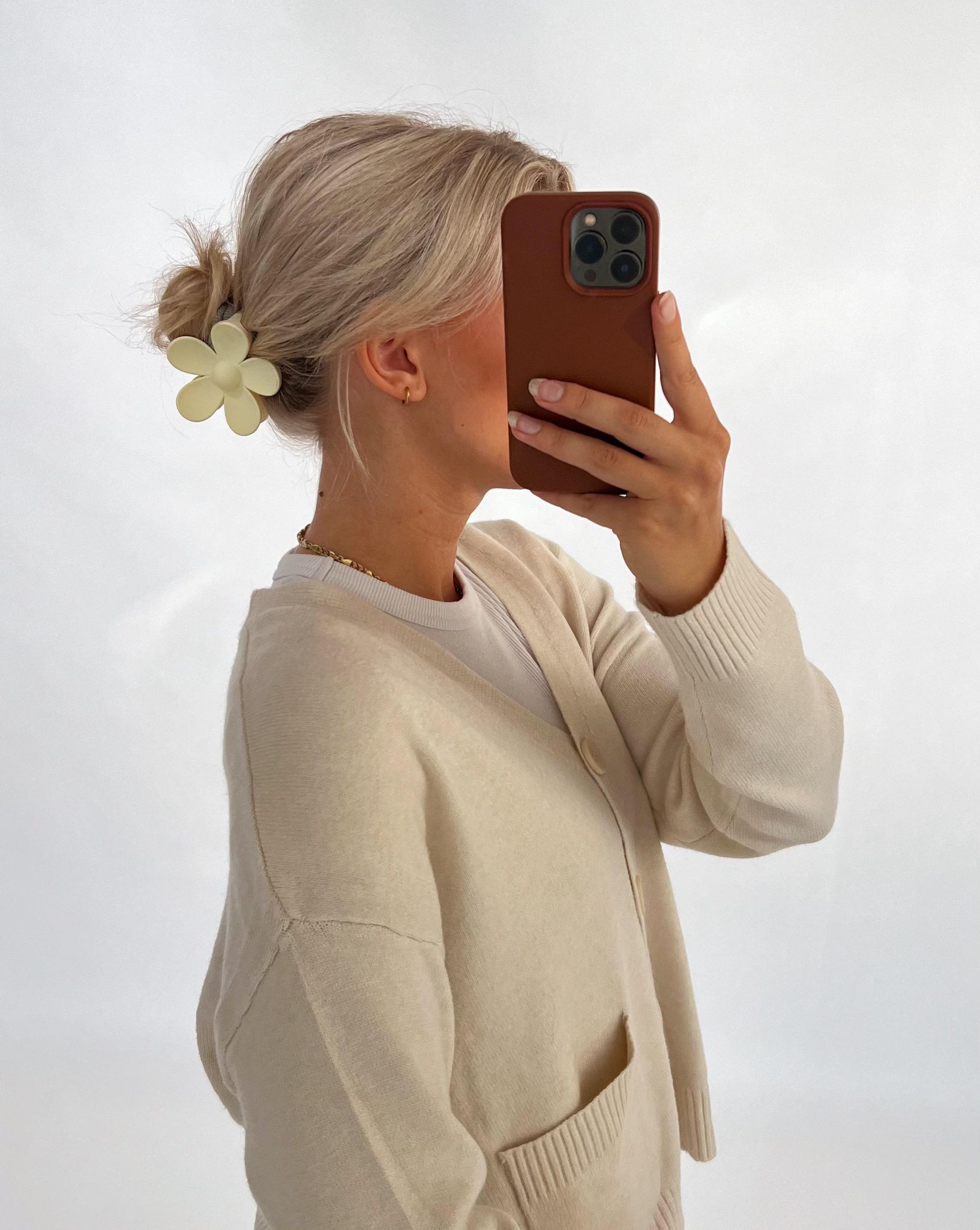 En person med lyst hår i en opsat frisure med en blomsterformet hårklemme tager et selfie med en mobiltelefon. Personen bærer en lys cardigan i et neutralt rum.