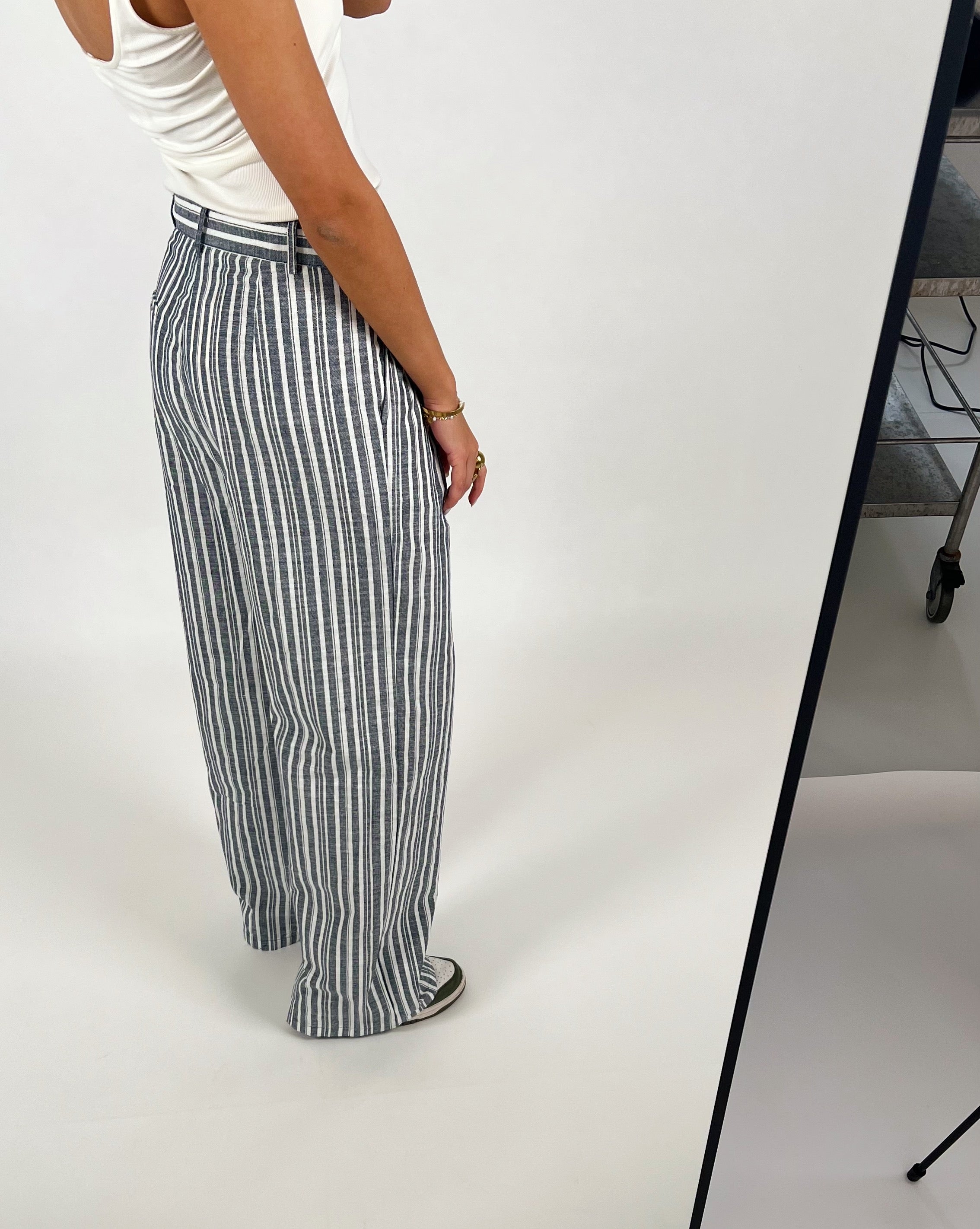 En person iført hvid tanktop og blå-hvid stribede bukser står mod en lys baggrund, mens de poserer i et fotostudie med hjul under en bordkant.
