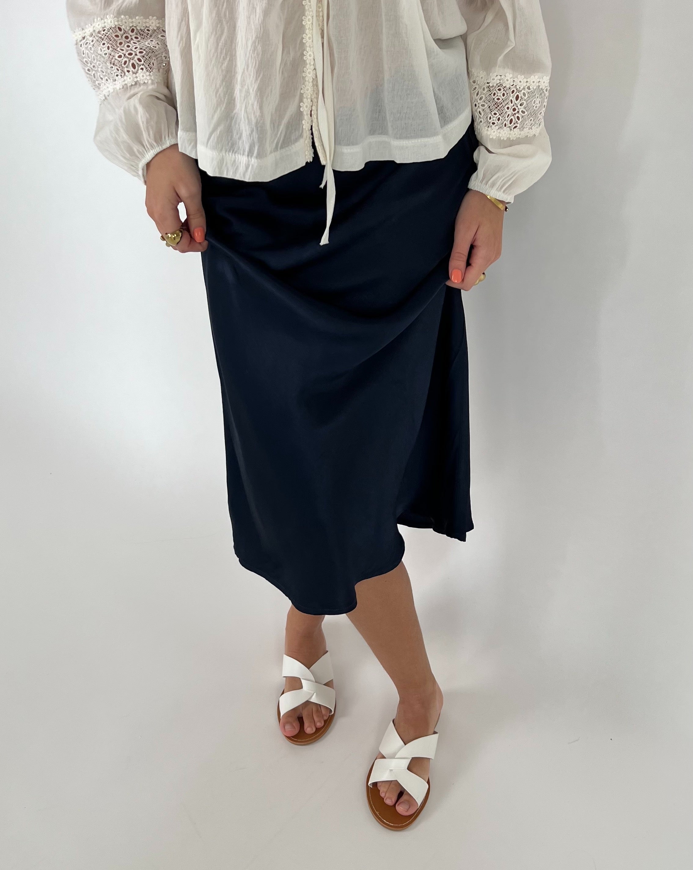 En person iført en hvid bluse med blonder holder i en mørk nederdel. De bærer hvide sandaler, mens de står i et neutralt, lyst rum.