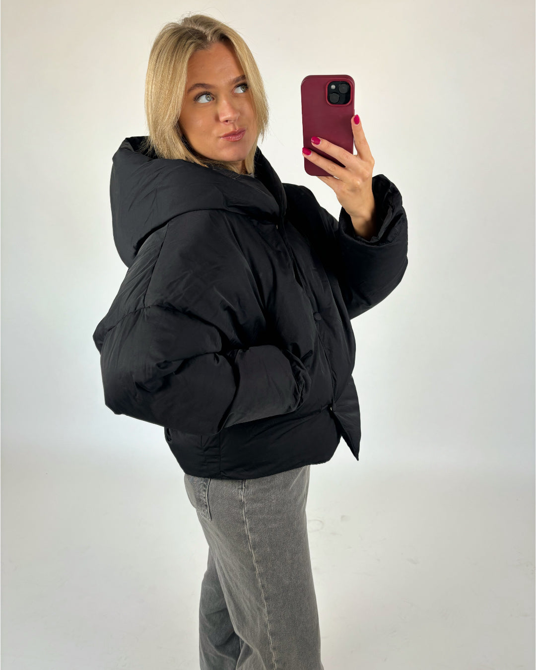 En person iført en sort dunjakke tager en selfie med en rød mobiltelefon. Personen står i et lys rum med en neutral baggrund.
