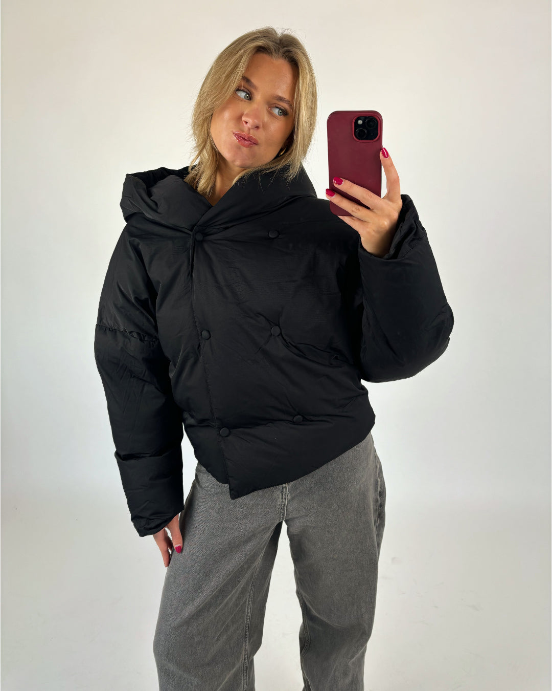 En person tager en selfie iført en sort, polstret jakke og grå bukser mod en hvid baggrund, mens de holder en mobiltelefon i en mørkerød etui.