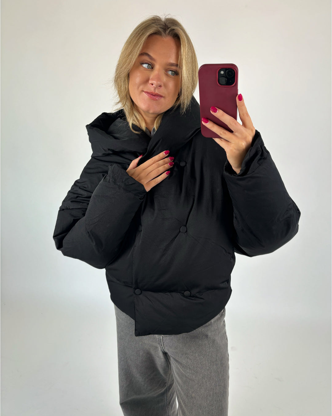 En kvinde bærer en sort, polstret jakke, mens hun tager en selfie med en rød telefon. Hun står i et neutralt oplyst rum, klædt i grå bukser.