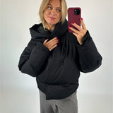 En kvinde bærer en sort, polstret jakke, mens hun tager en selfie med en rød telefon. Hun står i et neutralt oplyst rum, klædt i grå bukser.