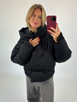 En kvinde bærer en sort, polstret jakke, mens hun tager en selfie med en rød telefon. Hun står i et neutralt oplyst rum, klædt i grå bukser.