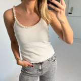 En person holder en telefon, tager et billede af sig selv iført en hvid top med perlekant og grå jeans. Baggrunden er enkel og lys.