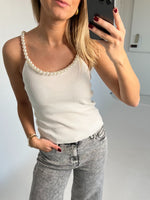 En person holder en telefon, tager et billede af sig selv iført en hvid top med perlekant og grå jeans. Baggrunden er enkel og lys.