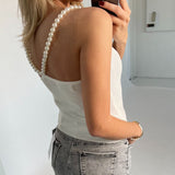 En kvinde fotograferer sig selv iført en hvid top med perleremme og grå jeans. Hun står i et lyst rum med en hvid baggrund og holder en telefon.