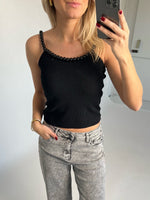 Kvinde iført sort top med perlepynt tager selfie. Hun har lyse jeans og rød neglelak. Baggrund er et lyst indendørs rum.