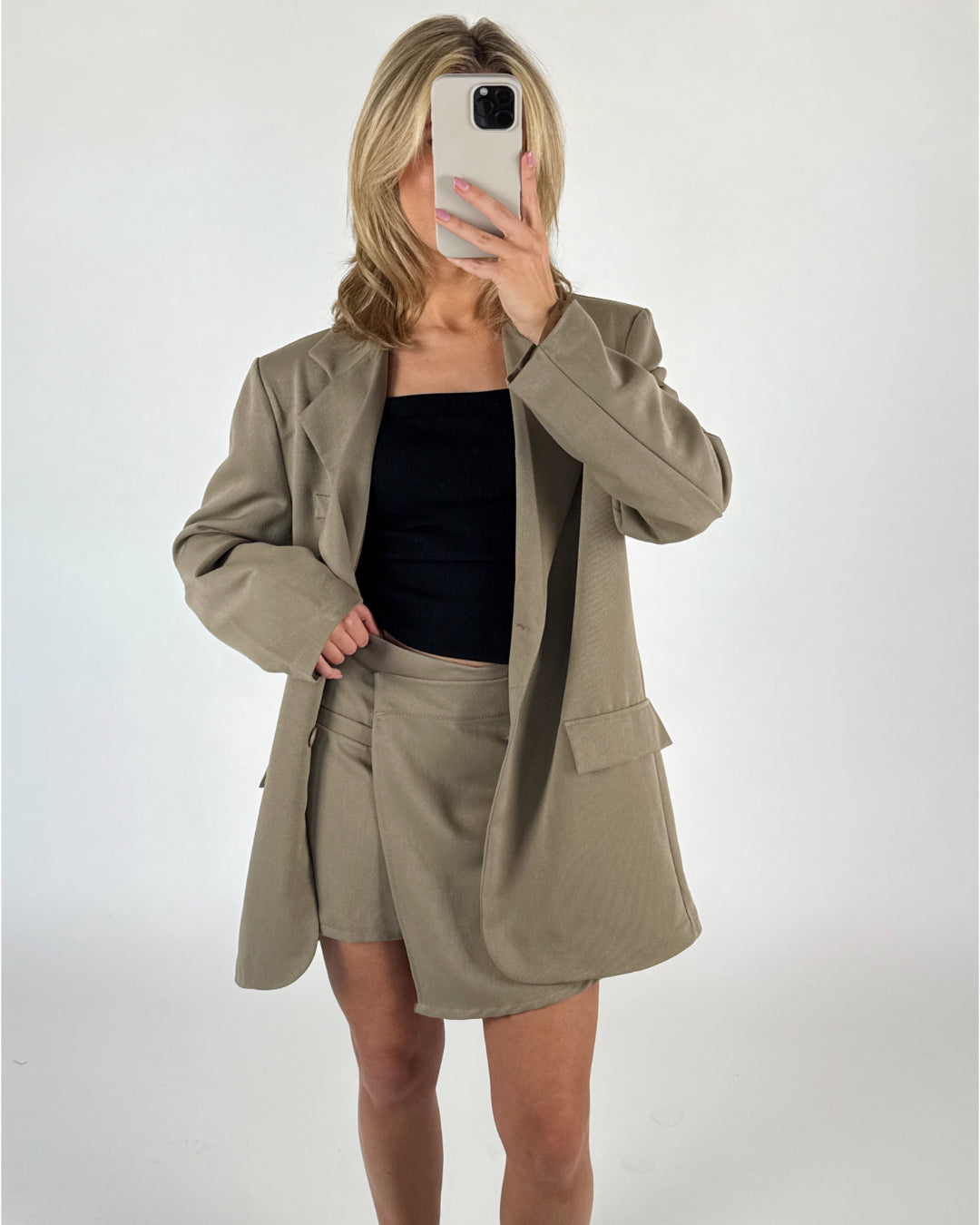 En person iført en beige blazer og nederdel tager et spejlbillede med sin smartphone. De står foran en hvid baggrund.
