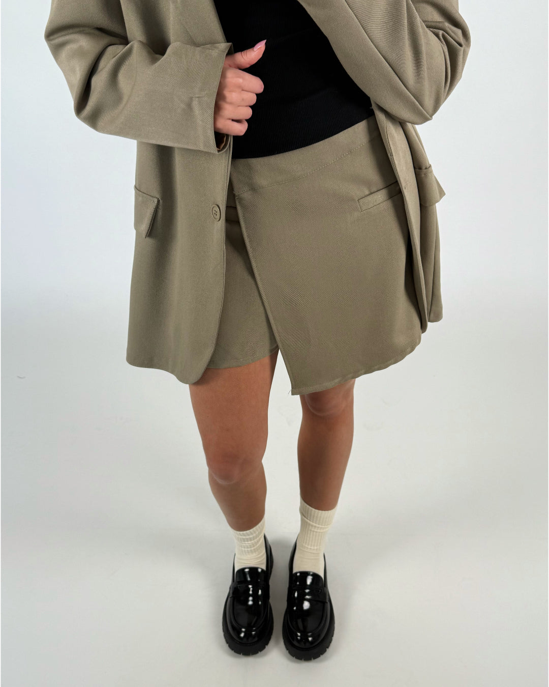 En person iført en beige jakke og nederdel står med krydsede hænder, iført sorte loafers og cremefarvede sokker, på en lys baggrund.