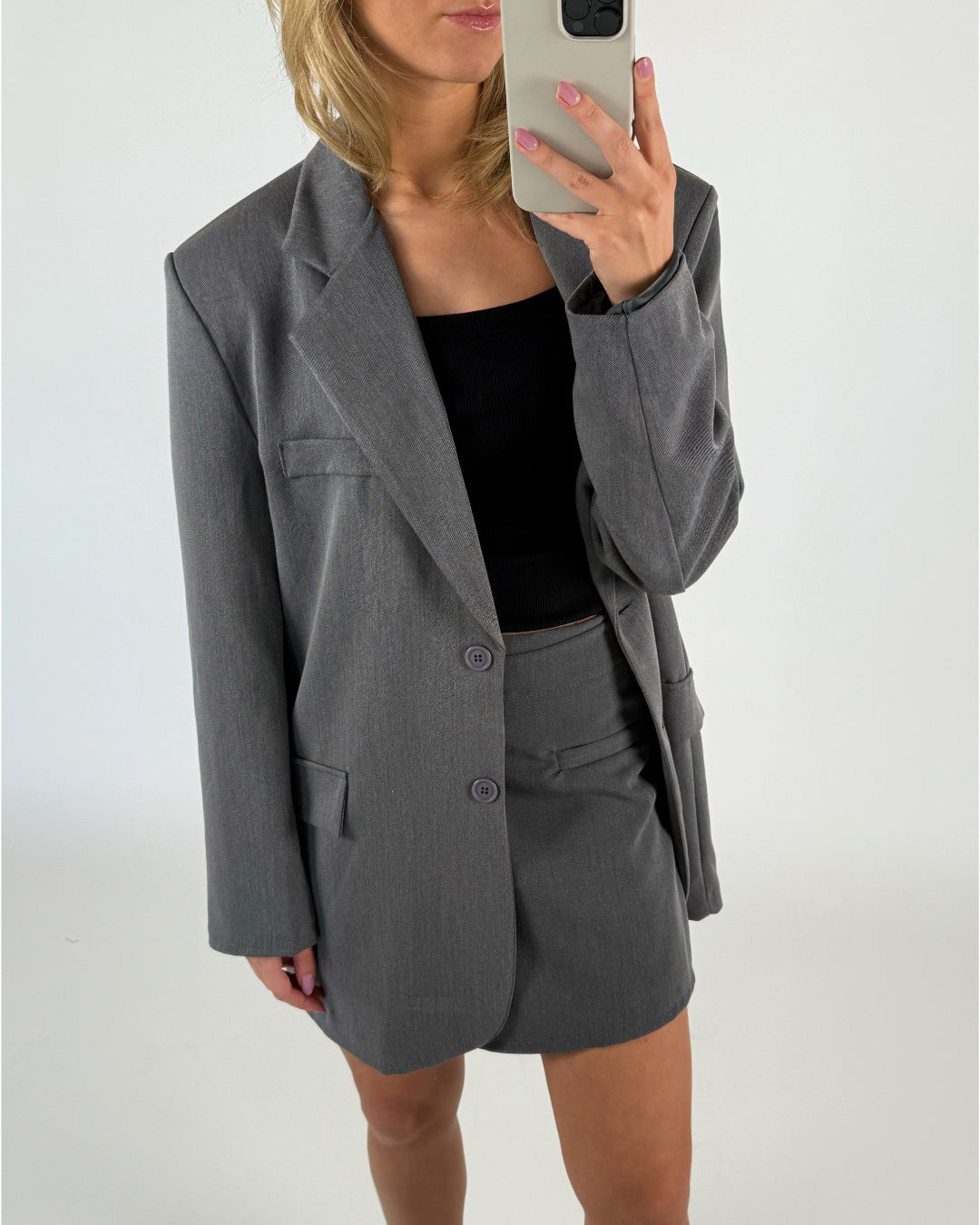 En kvinde iført en grå blazer og nederdel tager et spejlbillede med en smartphone i et neutralt, lys rum.
