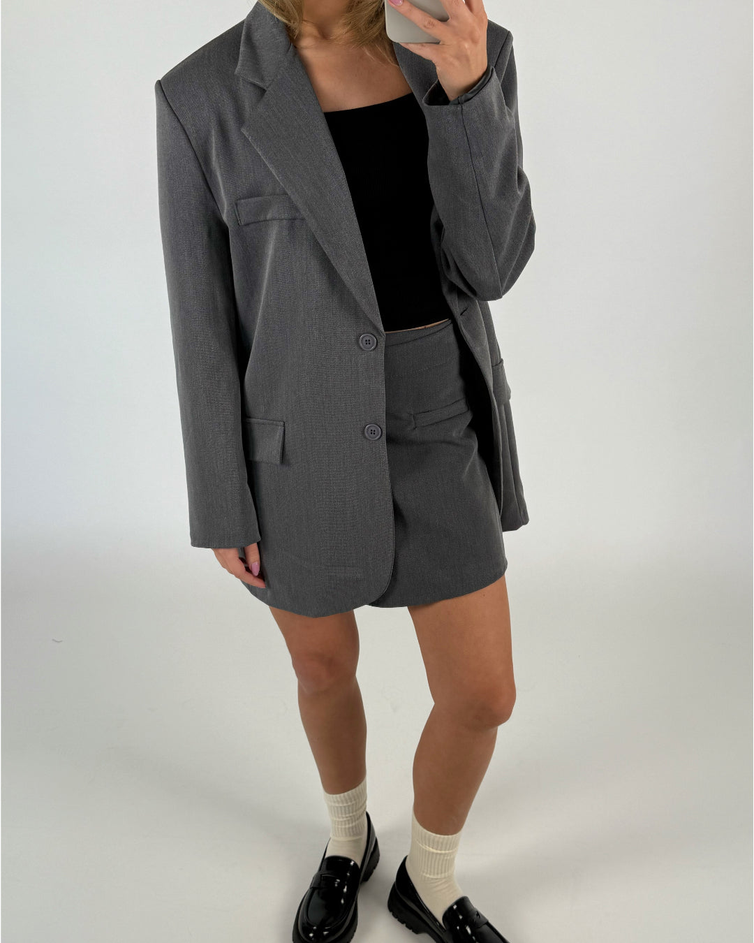 En person iført en grå blazer og nederdel står med en mobiltelefon foran ansigtet. De har sorte loafers og hvide sokker på. Baggrunden er minimalistisk, hvid.