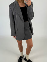 En person iført en grå blazer og nederdel står med en mobiltelefon foran ansigtet. De har sorte loafers og hvide sokker på. Baggrunden er minimalistisk, hvid.