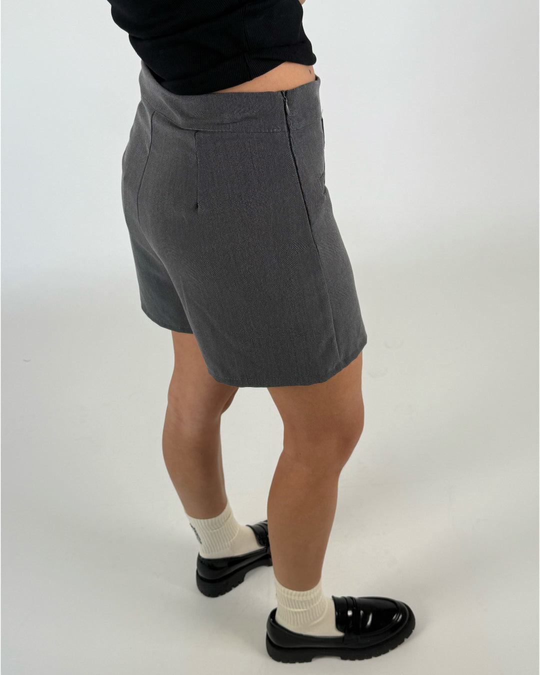 Person iført grå shorts står afslappet mod en lys baggrund. De bærer sorte sko og hvide sokker, mens en sort trøje dækker overkroppen.