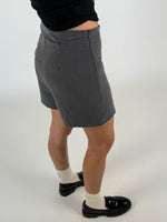 Person iført grå shorts står afslappet mod en lys baggrund. De bærer sorte sko og hvide sokker, mens en sort trøje dækker overkroppen.