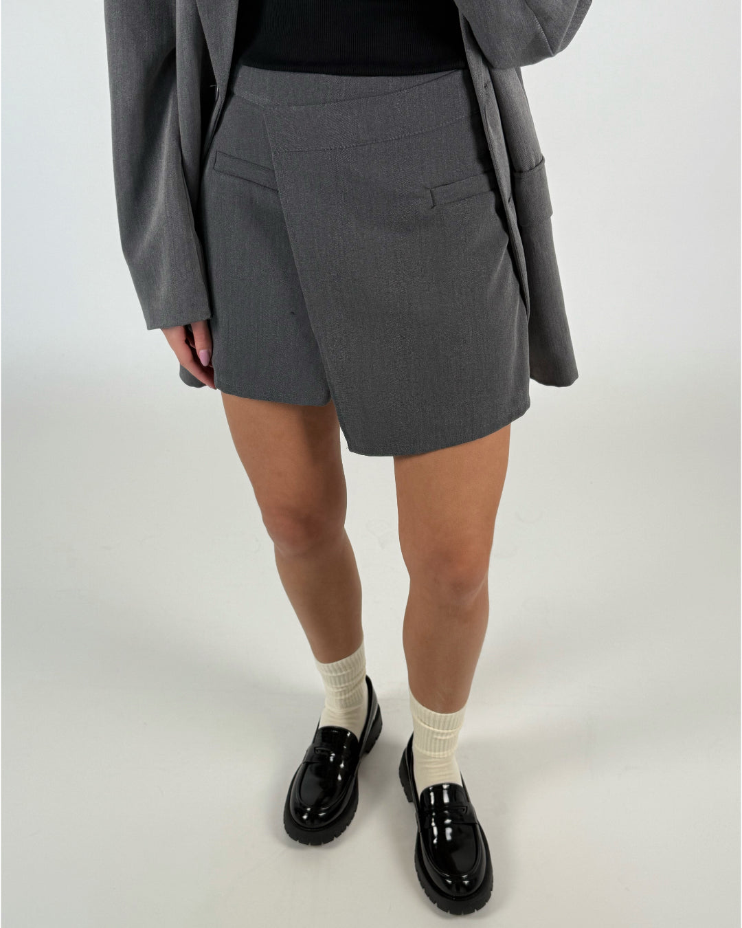En person iført grå shorts står, iført sorte laksko og cremefarvede sokker. En grå blazer hænger over den sorte top i et neutralt oplyst rum.