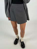 En person iført grå shorts står, iført sorte laksko og cremefarvede sokker. En grå blazer hænger over den sorte top i et neutralt oplyst rum.