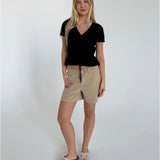 Kvinde står afslappet, iført sort bluse og beige shorts med sløjfesandaler, i et rum med hvid baggrund. Hun har langt lyst hår og smiler let.