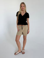 Kvinde står afslappet, iført sort bluse og beige shorts med sløjfesandaler, i et rum med hvid baggrund. Hun har langt lyst hår og smiler let.