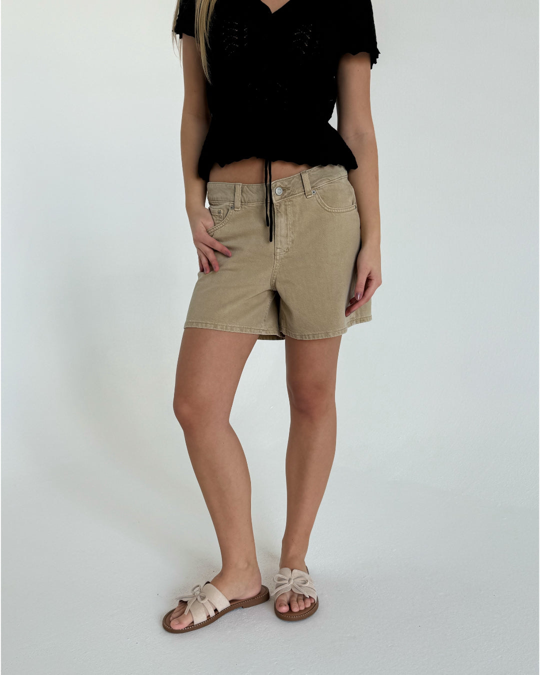 En person står iført beige shorts og sorte top i et lyst rum. De har en hånd i lommen og sandaler på fødderne.