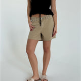 En person står iført beige shorts og sorte top i et lyst rum. De har en hånd i lommen og sandaler på fødderne.