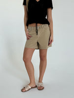 En person står iført beige shorts og sorte top i et lyst rum. De har en hånd i lommen og sandaler på fødderne.