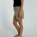 En person iført en sort bluse og beige shorts står i profil, iført lyse sandaler med sløjfer. Baggrunden er en ensfarvet, lys væg.