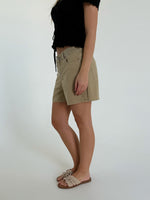En person iført en sort bluse og beige shorts står i profil, iført lyse sandaler med sløjfer. Baggrunden er en ensfarvet, lys væg.